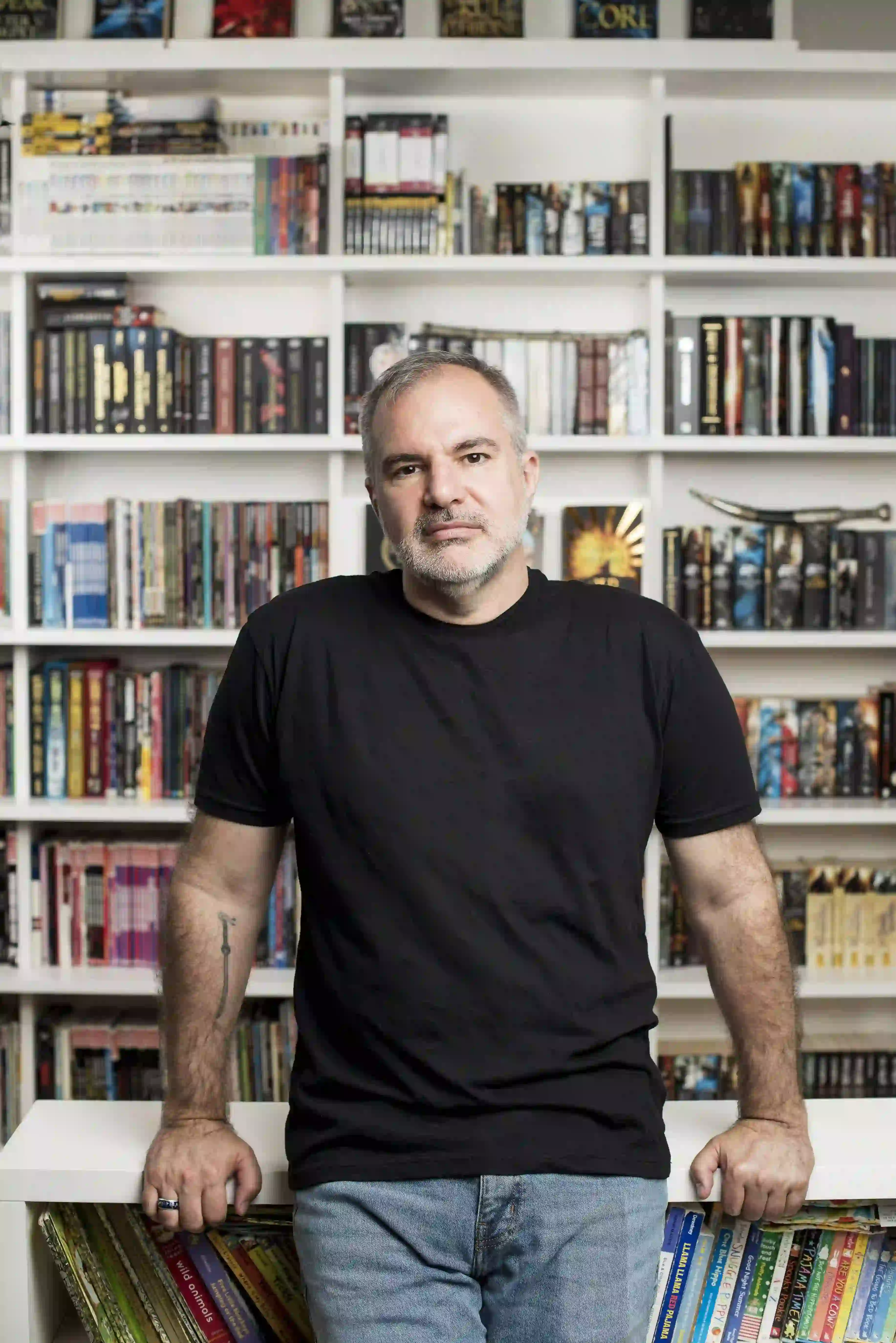 Pisarz Peter V. Brett stojący w czarnym t-shircie na tle domowej biblioteczki wypełnionej po brzegi książkami fantasy.